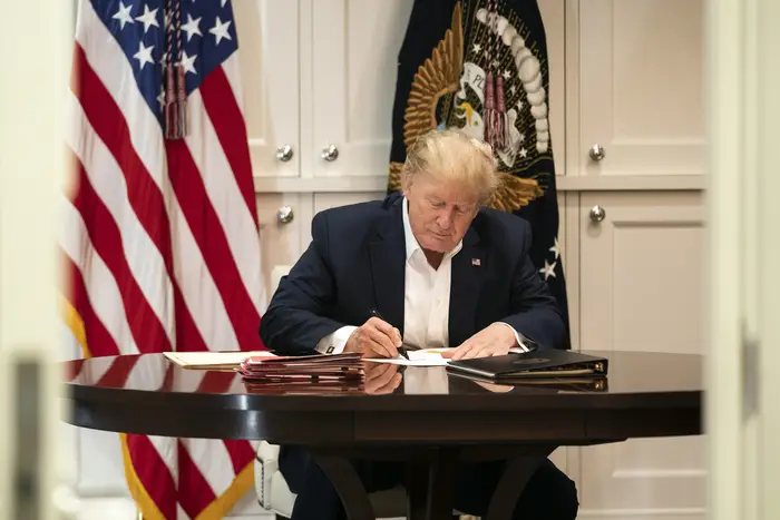 President Trump is seen in a shirt and suit jacket, no tie, sitting at a table signing a document, with white cabinetry behind him